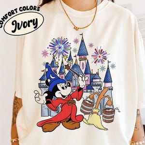 Könnte beinhalten: Elfenbeinfarbenes T-Shirt mit einem Cartoon von Mickey Mouse als Zauberer, der einen Zauberstab vor einem Schloss mit Feuerwerk hält. Der Text "COMFORT COLORS" und "Ivory" sind auf dem Shirt aufgedruckt.