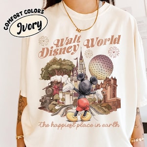 May include: Ivory-colored t-shirt with a vintage-style Walt Disney World graphic. The design features Mickey Mouse, a train, a castle, and the text "Walt Disney World" and "the happiest place on earth". The shirt has "Comfort Colors" printed on it.