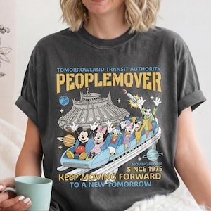 May include: Dark gray t-shirt featuring a Tomorrowland PeopleMover design with Mickey Mouse, Minnie Mouse, Donald Duck, and Goofy. The shirt includes the text "TOMORROWLAND TRANSIT AUTHORITY PEOPLEMOVER" and "KEEP MOVING FORWARD TO A NEW TOMORROW."