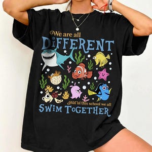 Op de afbeelding: Zwart t-shirt met een kleurrijke afbeelding van cartoonvissen en de tekst "We are all DIFFERENT, But in this school we all SWIM TOGETHER." Het ontwerp bevat een haai, een clownvis en andere zeedieren.