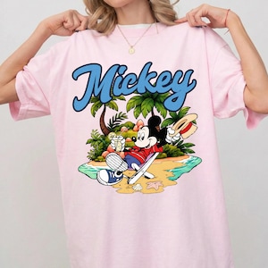 May include: Pink t-shirt featuring the word "Mickey" in blue script above a cartoon illustration of Mickey Mouse relaxing on a beach. Mickey is wearing a red shirt, blue shorts, and a straw hat, holding a drink.