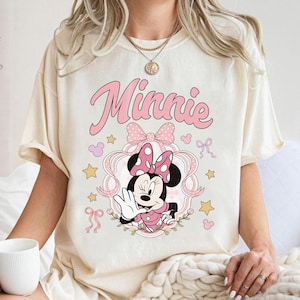 May include: A cream-colored t-shirt featuring a graphic of Minnie Mouse in a pink polka-dot bow and dress. The design includes the text Minnie in pink script, surrounded by pink ribbons, small yellow stars, and tiny pink mouse-ear silhouettes.