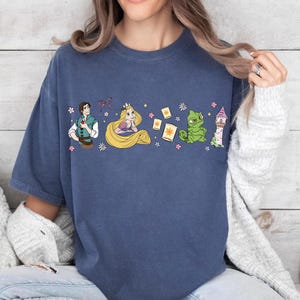 May include: A navy blue t-shirt featuring a colorful graphic of characters from a fairy tale. The design includes a prince, a princess with long blonde hair, a green frog, a tower, and floating lanterns. The shirt is made of a soft material.
