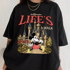 Puede incluir: Camiseta negra con un gr&aacute;fico de Mickey Mouse caminando frente a un castillo, con el texto "LIFE'S A WALK IN THE PARK" y "Walt Disney World" arriba. El dise&ntilde;o es en rojo, amarillo y beige.