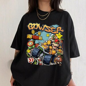 以下が含まれることがあります： 黒いTシャツ。カートに乗ったクッパのカラフルなグラフィック。オレンジと黄色の「Bowser」の文字。コイン、星、クエスチョンマークブロックなど、ビデオゲームを連想させるデザイン。