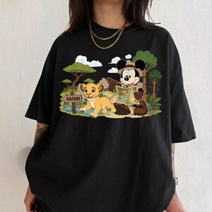 Op de afbeelding: Zwart t-shirt met een kleurrijke afbeelding van Mickey Mouse met een safarihoed, een kaart vasthoudend, en Simba het leeuwenjong. De sc&egrave;ne speelt zich af in een jungle met een "Safari"-bord.
