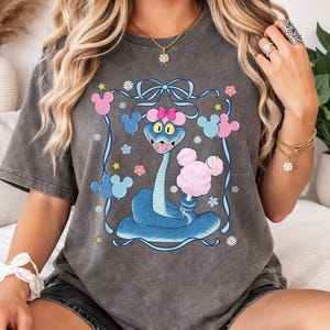 May include: Dark gray t-shirt featuring a cartoon snake with a blue body, holding pink cotton candy. The design includes pink and blue Mickey Mouse-shaped balloons, flowers, and a bow. The shirt is a unisex style.