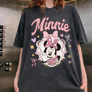 May include: A charcoal gray short-sleeve t-shirt featuring a graphic of Minnie Mouse winking and waving. The design includes the text "Minnie" in pink script, a large pink polka-dot bow, gold stars, and small pink ribbons on a dark background.