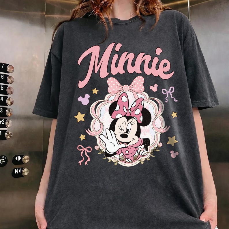 May include: A charcoal gray short-sleeve t-shirt featuring a graphic of Minnie Mouse winking and waving. The design includes the text "Minnie" in pink script, a large pink polka-dot bow, gold stars, and small pink ribbons on a dark background.