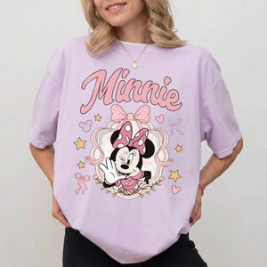 May include: A light purple t-shirt featuring a graphic of Minnie Mouse wearing a polka dot bow. The text Minnie is printed in pink above the character, surrounded by pink ribbons, yellow stars, and small Mickey Mouse head silhouettes.