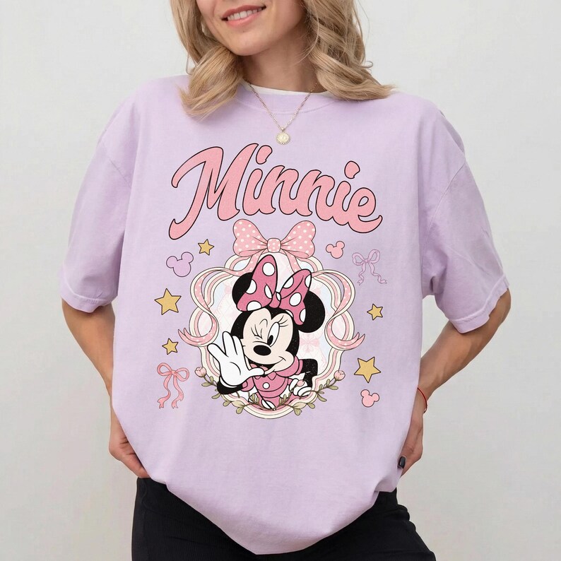 May include: A light purple t-shirt featuring a graphic of Minnie Mouse wearing a polka dot bow. The text Minnie is printed in pink above the character, surrounded by pink ribbons, yellow stars, and small Mickey Mouse head silhouettes.
