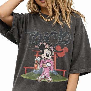 Peut inclure: T-shirt gris fonc&eacute; surdimensionn&eacute; avec un motif de Minnie Mouse en kimono rose, tenant une lanterne, avec le mot "Tokyo" au-dessus. Le design comprend des &eacute;l&eacute;ments japonais et un torii rouge.