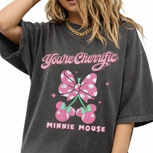 Pode incluir: Camiseta cinza escuro com la&ccedil;o rosa de bolinhas e design de cereja com orelhas da Minnie Mouse. O texto "You're Cherrific" e "MINNIE MOUSE" &eacute; impresso em rosa. A camiseta tem um visual vintage e desgastado.