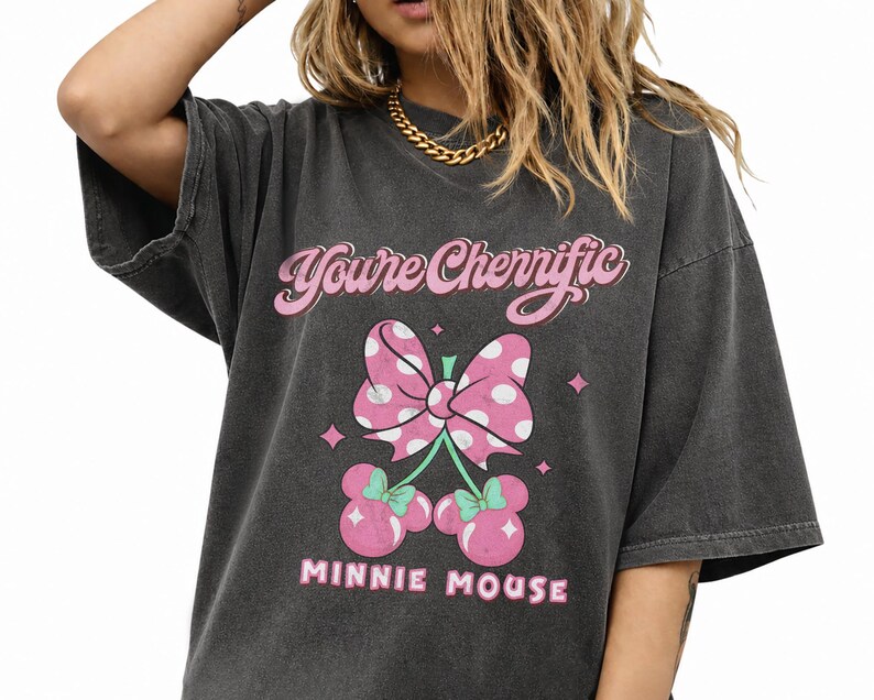 Pode incluir: Camiseta cinza escuro com la&ccedil;o rosa de bolinhas e design de cereja com orelhas da Minnie Mouse. O texto "You're Cherrific" e "MINNIE MOUSE" &eacute; impresso em rosa. A camiseta tem um visual vintage e desgastado.
