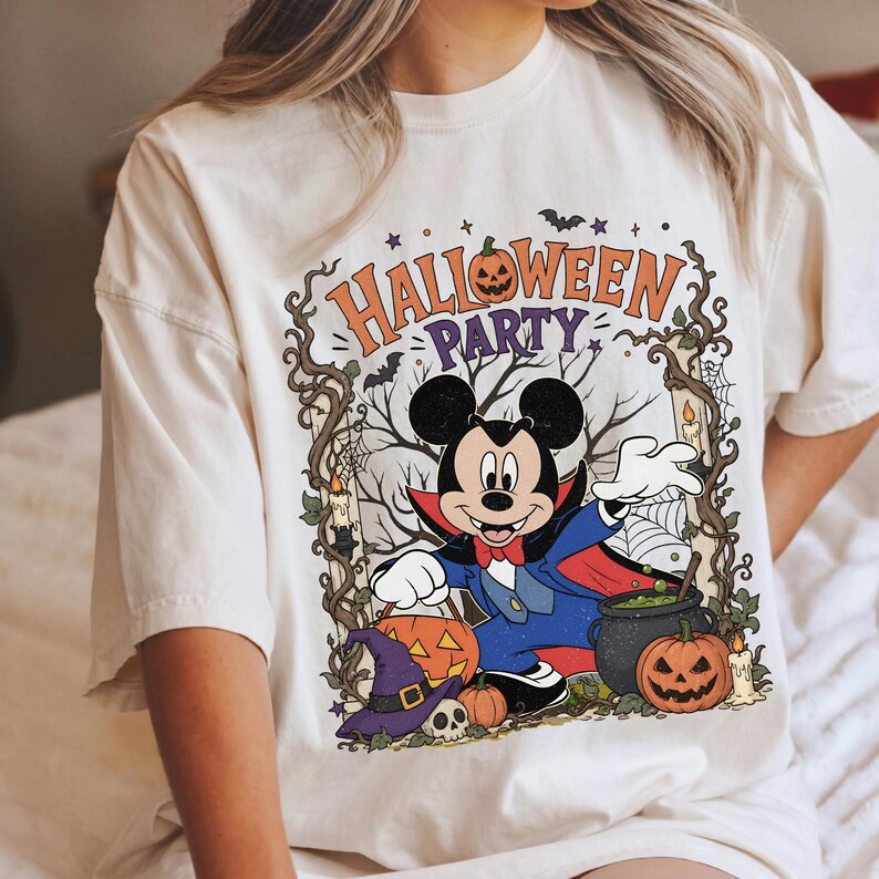 May include: A cream-colored t-shirt featuring a graphic of Mickey Mouse dressed as a vampire. The design includes the text HALLOWEEN PARTY, a jack-o'-lantern, a purple witch hat, a skull, a bubbling cauldron, and twisted tree branches.