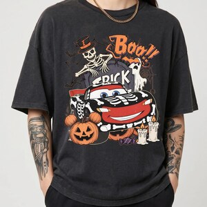 May include: A charcoal grey short-sleeve t-shirt featuring a graphic print of a race car decorated with skeleton bones, a skeleton wearing a top hat, a ghost, a jack-o'-lantern, candles, and the text "Boo!" and "TRICK" in orange and white.