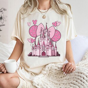May include: Cream-colored t-shirt featuring a pink castle graphic with a large bow and Mickey Mouse balloons. The castle has multiple turrets and a central arched doorway. The design is in a cartoon style, with varying shades of pink.