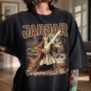 May include: Black t-shirt featuring a graphic of Jar Jar Binks holding a green lightsaber, with the text "JAR JAR" above and "Exsqueeze Me!" below. The design includes a battle scene with explosions and spaceships.