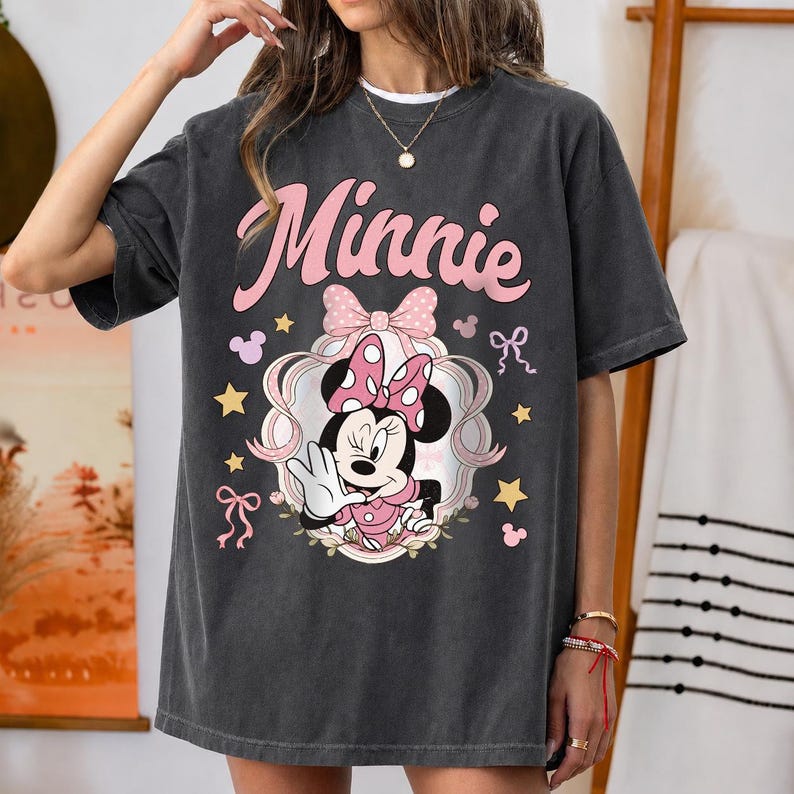 May include: A charcoal gray short-sleeve t-shirt featuring a graphic of Minnie Mouse in a pink polka-dot bow. The design includes the text Minnie in large pink script, surrounded by small yellow stars, pink bows, and mouse ear silhouettes.