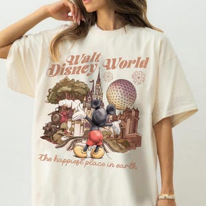 May include: Cream-colored t-shirt featuring a graphic of Mickey Mouse in front of a Disney World scene. The design includes a train, castle, and Spaceship Earth, with the text "Walt Disney World" and "The happiest place in earth".