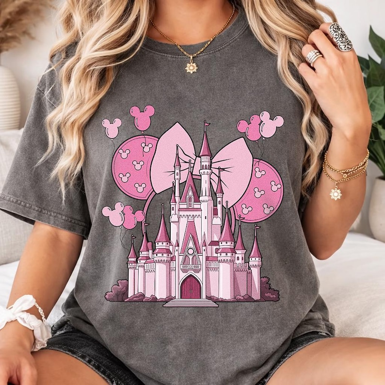 May include: Gray t-shirt featuring a pink castle design with a large bow and Mickey Mouse-shaped balloons. The castle has multiple turrets and a central entrance. The design is centered on the front of the shirt.