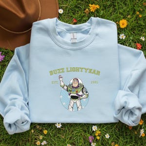 Puede incluir: Sudadera azul claro con un gr&aacute;fico de Buzz Lightyear y el texto "BUZZ LIGHTYEAR EST. 1995". El dise&ntilde;o presenta una imagen de dibujos animados del personaje en verde y blanco. La sudadera est&aacute; sobre un lecho de hierba verde.