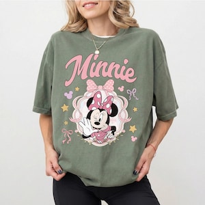 May include: An olive green short-sleeve t-shirt featuring a graphic print of Minnie Mouse in a pink polka-dot bow and dress. The design includes the text Minnie in pink script, surrounded by pink ribbons, small stars, and mouse-ear silhouettes.