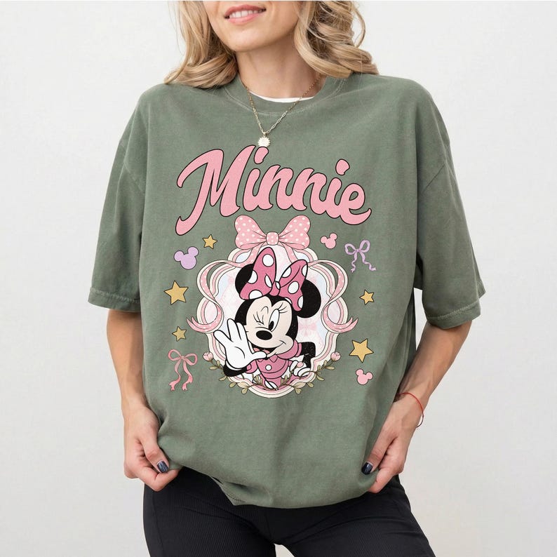 May include: An olive green short-sleeve t-shirt featuring a graphic print of Minnie Mouse in a pink polka-dot bow and dress. The design includes the text Minnie in pink script, surrounded by pink ribbons, small stars, and mouse-ear silhouettes.