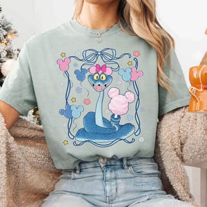 May include: A light green t-shirt featuring a cartoon snake with a pink bow, holding cotton candy. The snake is surrounded by Mickey Mouse-shaped balloons and flowers, all within a blue frame. The design is whimsical and colorful.