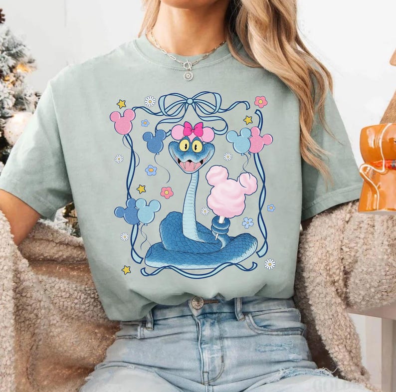 May include: A light green t-shirt featuring a cartoon snake with a pink bow, holding cotton candy. The snake is surrounded by Mickey Mouse-shaped balloons and flowers, all within a blue frame. The design is whimsical and colorful.