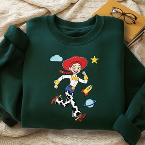 May include: Dark green sweatshirt with a cartoon character illustration. The design includes a character with a red hat, white shirt, and cow-print trousers, a rocket, planet, star, and cloud. The sweatshirt has a crew neck and long sleeves.