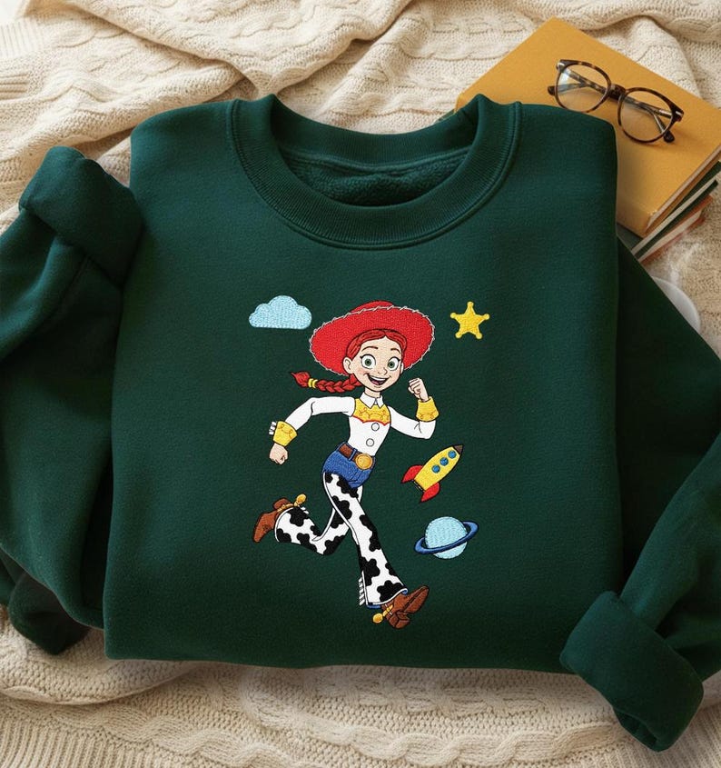 May include: Dark green sweatshirt with a cartoon character illustration. The design includes a character with a red hat, white shirt, and cow-print trousers, a rocket, planet, star, and cloud. The sweatshirt has a crew neck and long sleeves.