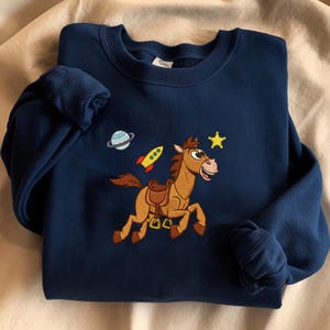 Pu&ograve; includere: Felpa blu navy con ricamo di un cavallo dei cartoni animati, un razzo, un pianeta e una stella. Il cavallo ha una sella. Il design &egrave; nei toni del marrone, rosso, giallo e blu. Girocollo e maniche arrotolate.