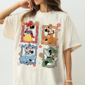 May include: Cream-colored t-shirt featuring four framed illustrations of Minnie Mouse in different outfits. The frames are in red, orange, blue, and green, with floral accents. Minnie is depicted in a Snow White-inspired dress, an orange gown, a blue dress, and a fairy costume.