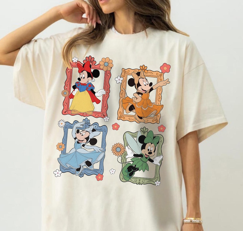 May include: Cream-colored t-shirt featuring four framed illustrations of Minnie Mouse in different outfits. The frames are in red, orange, blue, and green, with floral accents. Minnie is depicted in a Snow White-inspired dress, an orange gown, a blue dress, and a fairy costume.