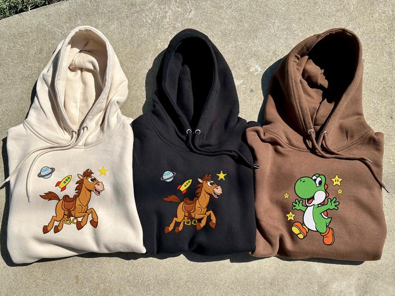 Pu&ograve; includere: Tre felpe con cappuccio beige, nere e marroni. Ognuna presenta una diversa grafica a cartoni animati: un cavallo con un razzo, un cavallo con un razzo e un personaggio di dinosauro verde. Le felpe hanno coulisse e una tasca frontale.