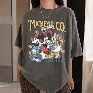May include: Dark gray t-shirt featuring a graphic of Mickey Mouse and friends, with the text "Mickey & Co. EST. 1928" in yellow. The design includes Mickey, Minnie, Donald Duck, Daisy Duck, Goofy, and Pluto.