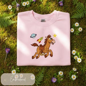 Pu&ograve; includere: Una felpa rosa chiaro con un ricamo di un cavallo dei cartoni animati, un razzo, un pianeta e una stella. La parola "Embroidered" &egrave; su un'etichetta. La felpa &egrave; mostrata su muschio verde con margherite e altri fiori.