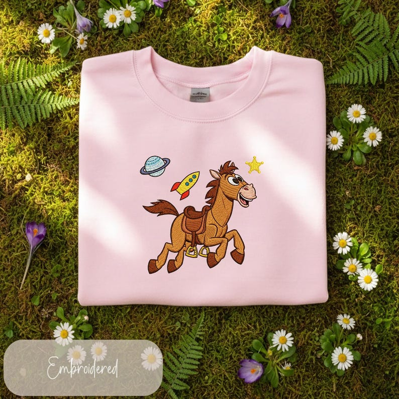 Pu&ograve; includere: Una felpa rosa chiaro con un ricamo di un cavallo dei cartoni animati, un razzo, un pianeta e una stella. La parola "Embroidered" &egrave; su un'etichetta. La felpa &egrave; mostrata su muschio verde con margherite e altri fiori.