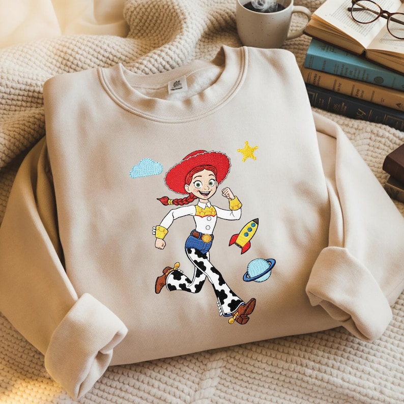May include: A beige sweatshirt with an embroidered cartoon character in a red hat and cow-print trousers. The design includes a rocket, a planet, a star, and a cloud. The character is running with a determined expression.
