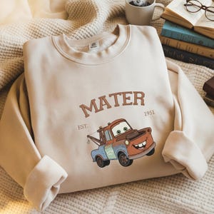 Può includere: Felpa girocollo beige con un'immagine a cartoni animati di Cricchetto, il testo sopra dice "MATER" e sotto "EST. 1951". La grafica è nei toni del marrone, rosso, blu e verde. La felpa è su una superficie strutturata.