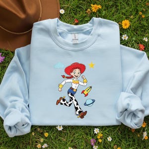 May include: Pale blue sweatshirt with a cartoon image of a running cowgirl in a red hat, white shirt, and cow-print trousers. A yellow star, rocket, and planet are also shown. The sweatshirt has long sleeves and a crew neck.