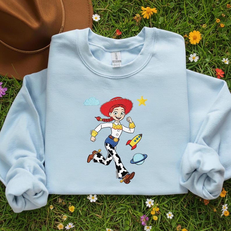 May include: Pale blue sweatshirt with a cartoon image of a running cowgirl in a red hat, white shirt, and cow-print trousers. A yellow star, rocket, and planet are also shown. The sweatshirt has long sleeves and a crew neck.