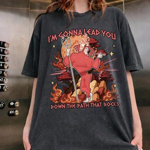 May include: Dark gray t-shirt with a graphic of a red devil holding a trident, standing in front of a fiery background. Text on the shirt reads "I'M GONNA LEAD YOU DOWN THE PATH THAT ROCKS."