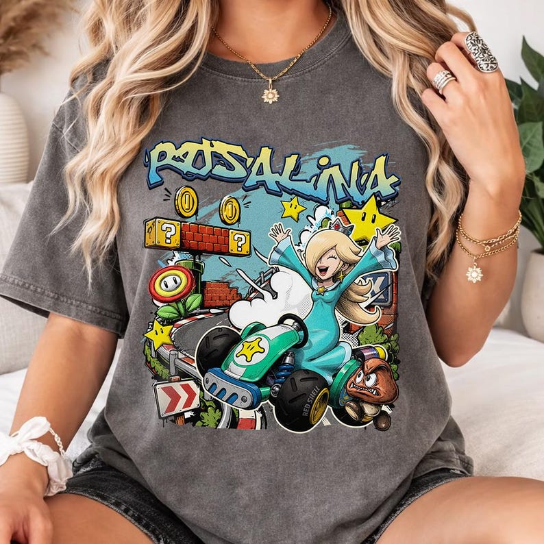 May include: Dark gray t-shirt featuring a colorful graphic of Rosalina from Mario Kart. The design includes the word "Rosalina" in graffiti style, coins, stars, and a kart. The shirt is a comfortable, casual style.