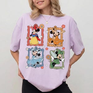 May include: A light purple t-shirt featuring four framed images of Minnie Mouse in different outfits. The frames are decorated with flowers. Minnie is dressed as Snow White, a princess, a ballerina, and a fairy.
