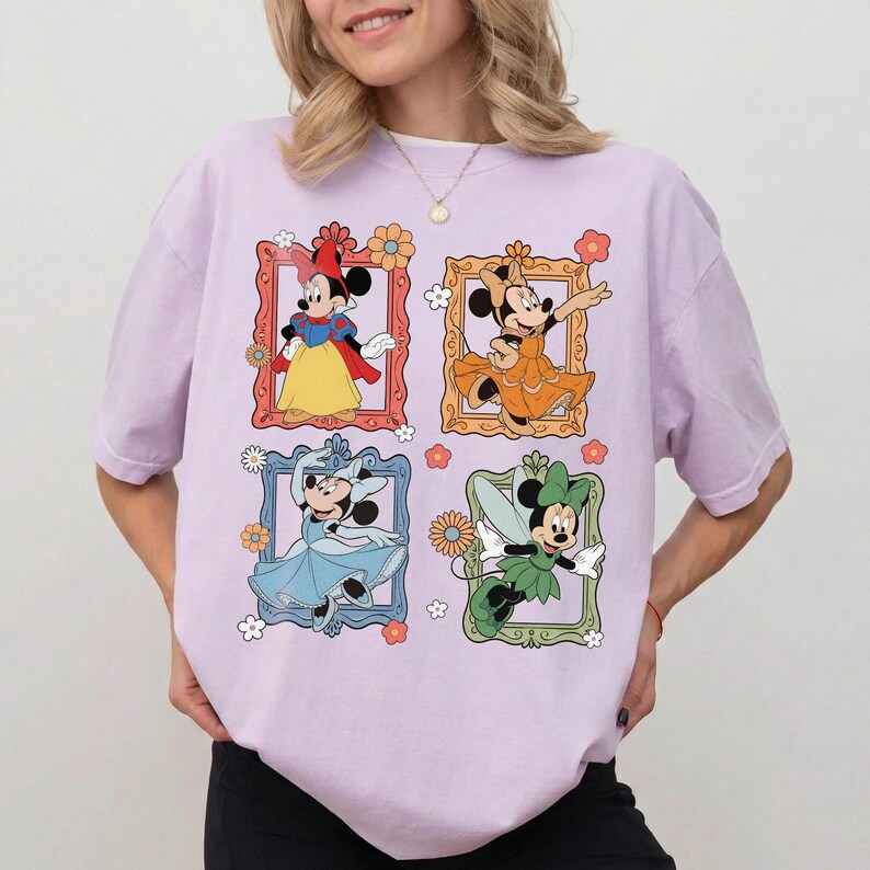 May include: A light purple t-shirt featuring four framed images of Minnie Mouse in different outfits. The frames are decorated with flowers. Minnie is dressed as Snow White, a princess, a ballerina, and a fairy.