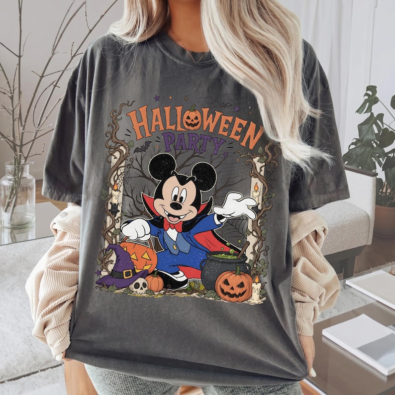 May include: A charcoal gray t-shirt featuring a graphic of Mickey Mouse dressed as a vampire. The design includes the text "HALLOWEEN PARTY" with pumpkins, a witch hat, a skull, and a cauldron set against a dark, spooky tree background.