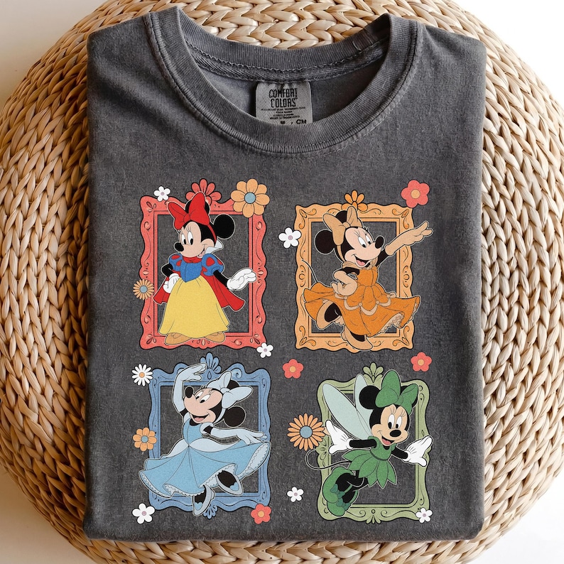 May include: Dark gray t-shirt featuring four framed Minnie Mouse illustrations in different outfits. The illustrations are in red, orange, blue, and green frames with floral accents. The shirt has a crew neck and the brand "Comfort Colors" is visible.