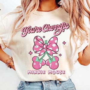 Pode incluir: Camiseta cor marfim com la&ccedil;o rosa de bolinhas brancas e design de cerejas. O texto "You're Cherrific" est&aacute; acima do la&ccedil;o e "Minnie Mouse" abaixo. A camiseta tem um corte folgado.